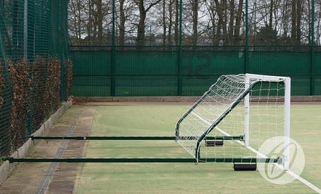 A Harrod Sport 4G Fence Folding Goal (3.66m x 1.22m, 3.5m to 5m projection) with wheels is lying on its side on an outdoor field next to a fenced area with trees in the background. This portable, folding 5-a-side goal is ideal for use on synthetic surface football pitches.