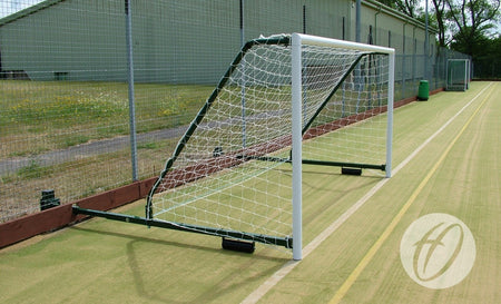 A Harrod Sport 4G Fence Folding Goal - 16 x 6, with a net attached, stands on an outdoor artificial turf field near a metal fence and building. Yellow field lines run alongside the goal.