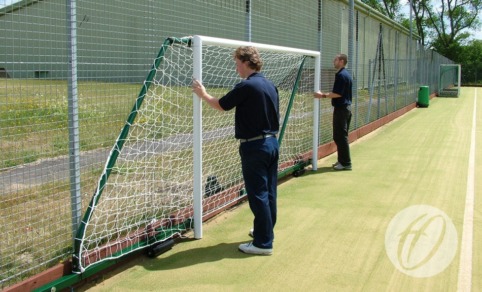 Two people are moving a Harrod Sport 4G Fence Folding Goal—7V7/5V5, with a projection between 3.5m and 5m—complete with net, across an artificial turf sports field next to a tall chain-link fence.