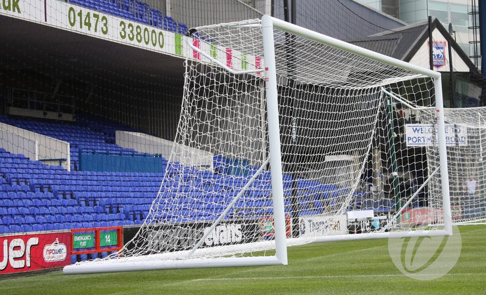 A Harrod Sport 3G Hinged Portagoal with a net stands on a green field in a stadium with blue seats; advertising boards and a building appear in the background.