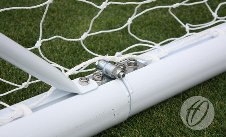 Close-up of a metal hinge and bolts securing the Harrod Sport 3G Hinged Portagoal's white aluminium post and net on green artificial turf. A circular logo with a stylized "f" appears in the bottom right corner.