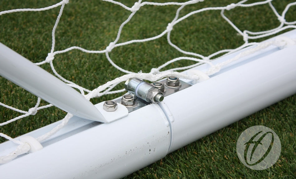 Close-up of a metal hinge and bolts securing the Harrod Sport 3G Hinged Portagoal's white aluminium post and net on green artificial turf. A circular logo with a stylized "f" appears in the bottom right corner.