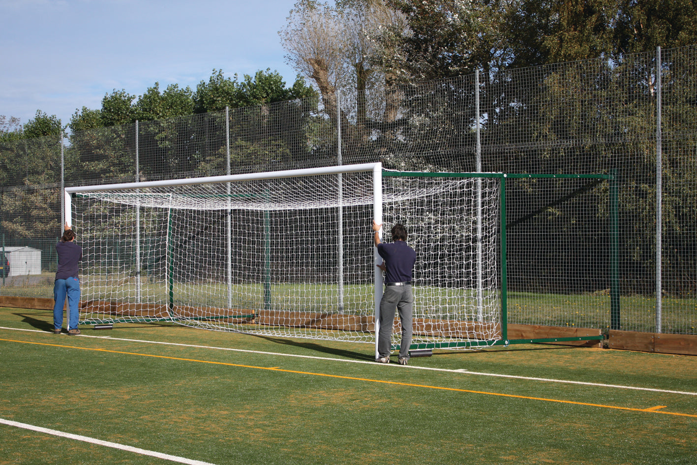 Two people move a Harrod Sport 3G Fence Folding Goal - Senior (2.3M to 3.5M projection) across an outdoor artificial turf field on a sunny day, with trees and a tall chain-link fence in the background.