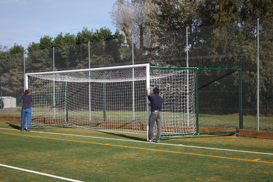 Two people move a Harrod Sport 3G Fence Folding Goal - Senior (2.3M to 3.5M projection) across an outdoor artificial turf field on a sunny day, with trees and a tall chain-link fence in the background.