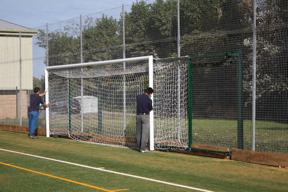 Two people are moving a Harrod Sport 3G Fence Folding Goal – Senior, 2.3m to 3.5m proj., near a wire fence at the edge of a sports field, with trees and a building visible in the background.