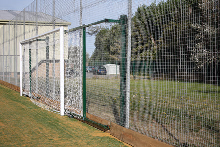 The Harrod Sport 3G Fence Folding Goal - Senior, 2.3M To 3.5M Proj. is set up beside a tall metal fence on artificial grass, with trees, parked cars, and a shed in the background.