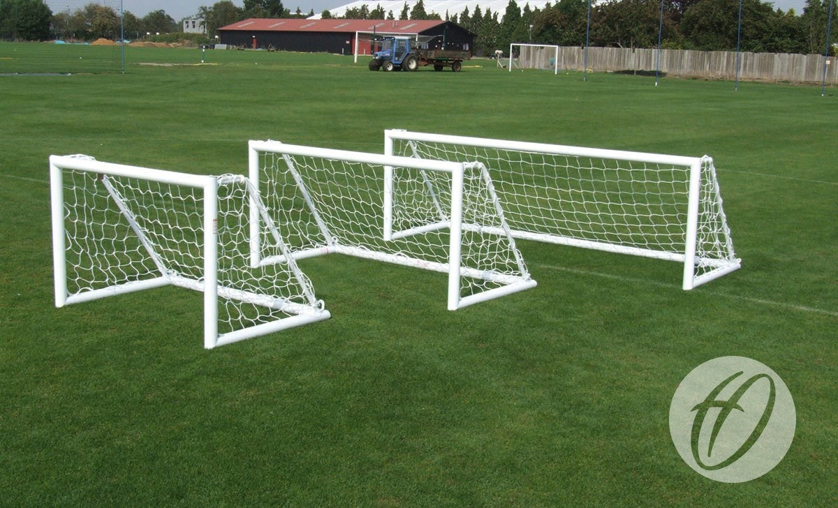 Three different-sized white soccer goals, including a mini training goal and a larger Harrod Sport Spare Target Goal Net (3m x 1m), are placed on a grassy field, with a building, tractor, and trees visible in the background. The image has a circular watermark in the bottom right corner.