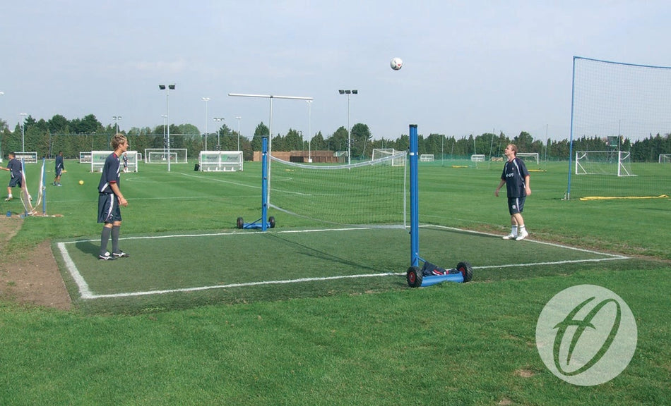 Two players enjoy a sunny match of foot tennis on grass with the Harrod Sport Premier Head Tennis unit, featuring an adjustable net. Soccer goals and the head tennis equipment make excellent training aids for sports clubs in the background.