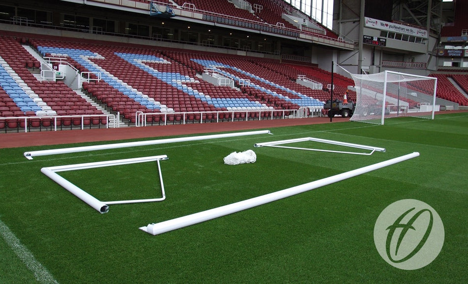 Disassembled Harrod Sport 3G Demountable Portagoal - Senior (7.32m x 2.44m) aluminium frame parts lie on the green field in a stadium with empty red and blue seats in the background.