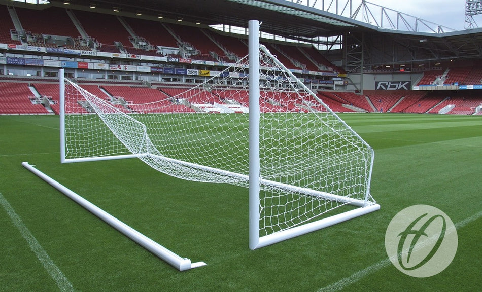 A Harrod Sport 3G Demountable Portagoal - Senior (7.32m x 2.44m) made of white aluminium lies tipped over on its side on a green soccer field inside an empty stadium with red seats, while advertising boards line the stands in the background.