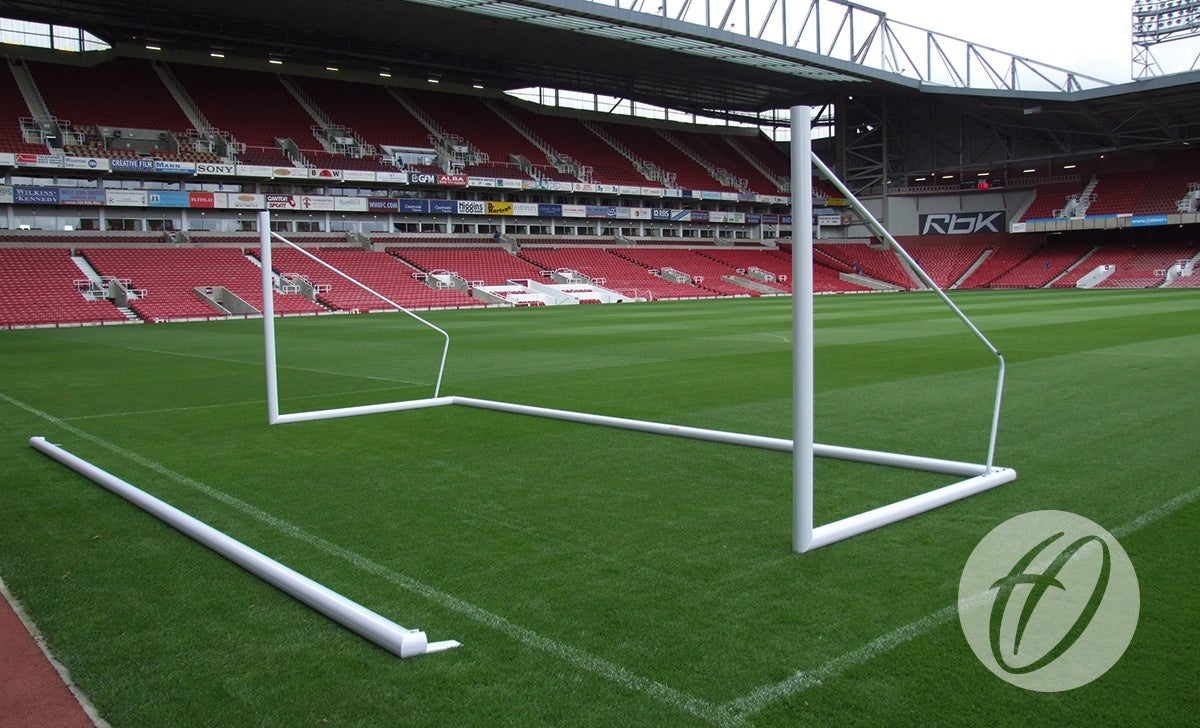 A partially assembled Harrod Sport 3G Demountable Portagoal - Senior (7.32M x 2.44M) white soccer goal frame rests on the green field of an empty stadium with red seats, with some stadium logos and seating visible in the background.