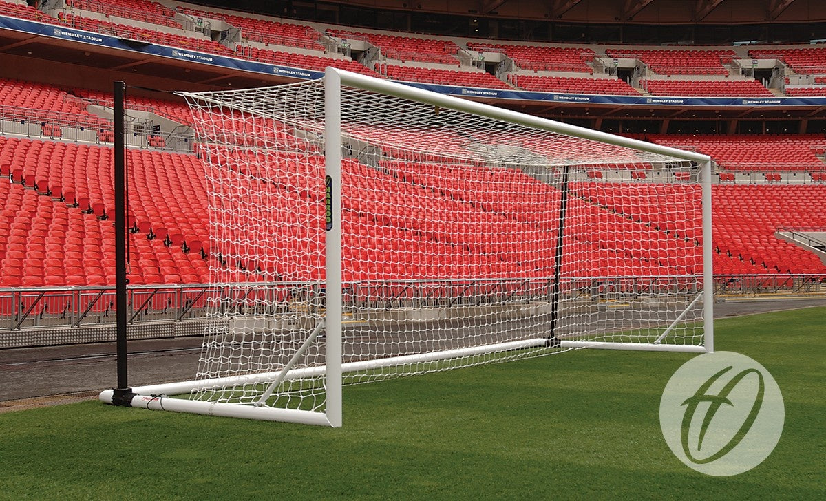 A Harrod Sport 3G Foldaway Euro Portagoal with a net is set on green grass in an empty stadium with red seats. A circular logo featuring a stylized H appears in the lower right corner.