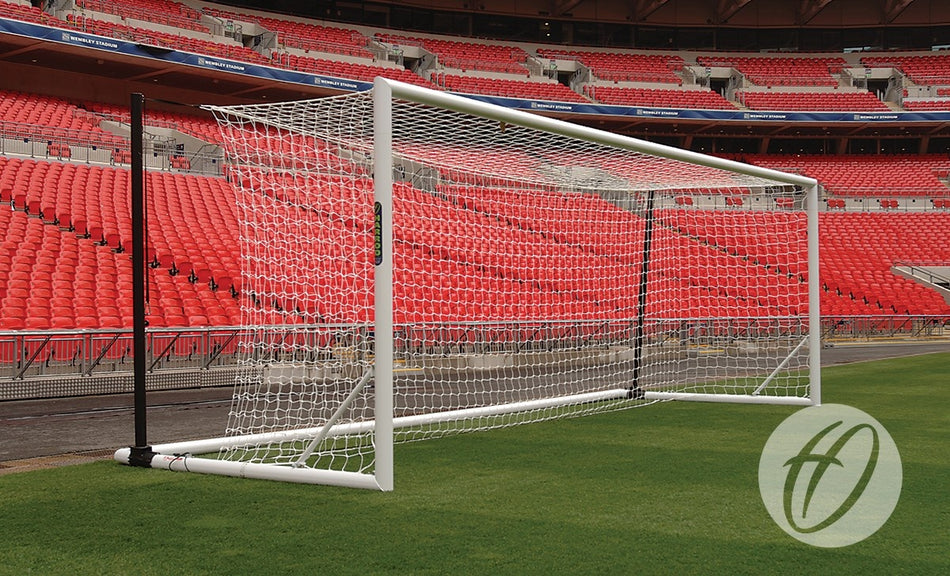 A Harrod Sport 3G Foldaway Euro Portagoal with a net is set on green grass in an empty stadium with red seats. A circular logo featuring a stylized H appears in the lower right corner.