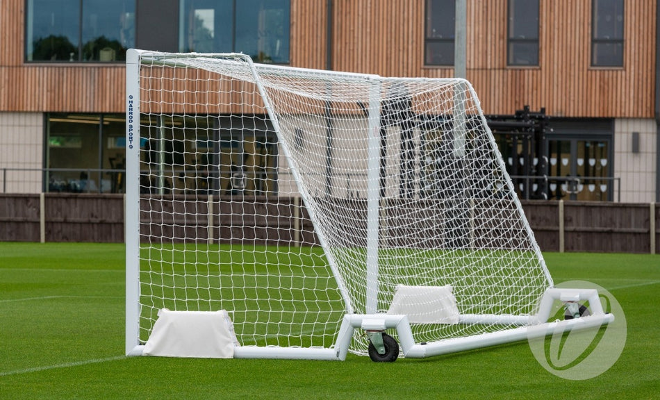 A Harrod Sport Harrod 4G Swivel Senior Portagoal with a net and wheels stands on a well-kept grass field, perfect for Premier League coaches. A modern building with large windows can be seen in the background.
