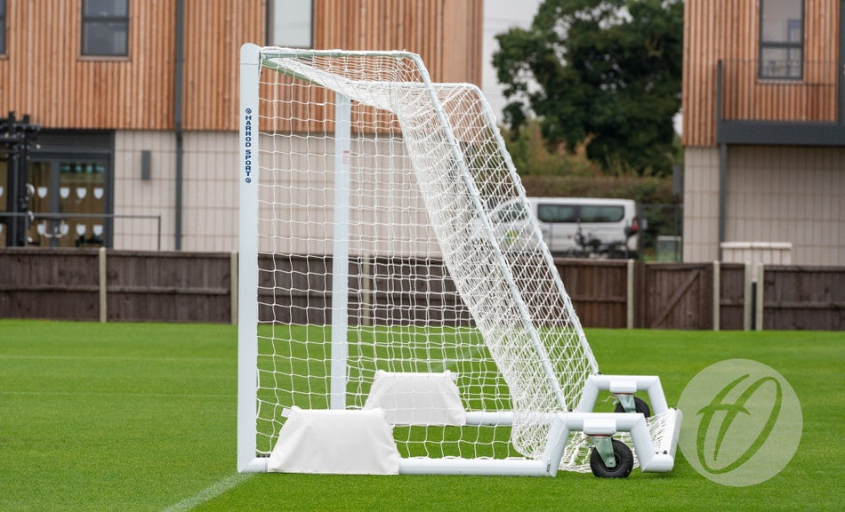 A Harrod Sport 4G Swivel Weighted 9V9 Portagoal with wheels on its base stands on green grass before a fence, with a van, trees, and modern buildings in the background—ideal for training or as a versatile 4G Portagoal.