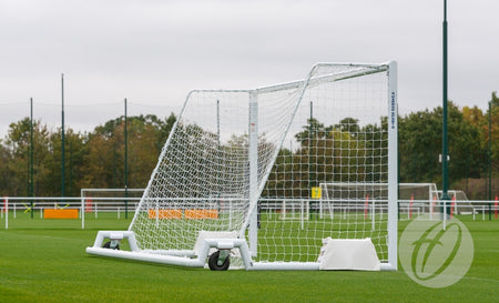 The Harrod Sport 4G Swivel 7v7/5v5 Portagoal, a white aluminium training goal with wheels, stands on a green field bordered by white fencing and trees beneath an overcast sky.
