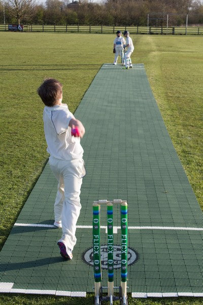 A young cricket player bowls toward a batter, both in white, on a green Flicx UK 2G Flicx Match Pitch outdoors, with stumps and bails clearly visible in the foreground.