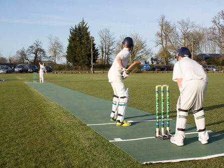 Three boys in cricket gear play outdoors on a green 2G Flicx Match Pitch by Flicx UK; one is bowling, another bats, and the third keeps wicket, with trees and parked cars in the background.