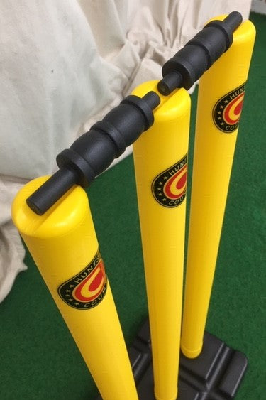 The Hunts County Flik Cricket Stumps & Base Set (Yellow) features three bright yellow stumps with black bails and a sturdy base, all bearing the Hunts County logo, standing on green turf with a white curtain in the background.