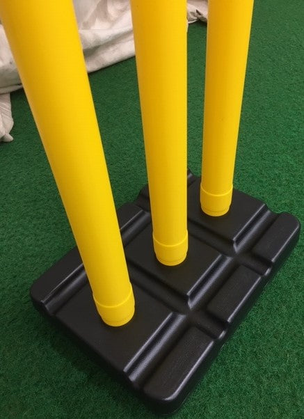 The Hunts County Flik Cricket Stumps & Base Set (Yellow) features three yellow plastic stumps standing in a black base by Hunts County, set on green artificial turf.