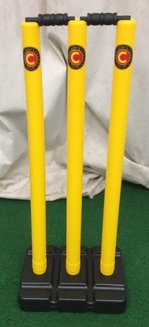 The Hunts County Flik Cricket Stumps & Base Set (Yellow) by Hunts County features three yellow plastic stumps with a black base and black bails, arranged on green turf against a light-colored fabric backdrop.