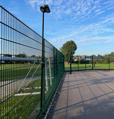 A Ritelite Sports Lite Fence Mast Adapter is mounted to a tall metal fence with a camera and height-adjustable mast, separating a paved sports court from a grassy field under a blue sky. One person stands on the court in the distance.
