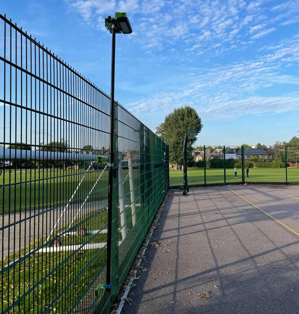 A Ritelite Sports Lite Fence Mast Adapter is mounted to a tall metal fence with a camera and height-adjustable mast, separating a paved sports court from a grassy field under a blue sky. One person stands on the court in the distance.