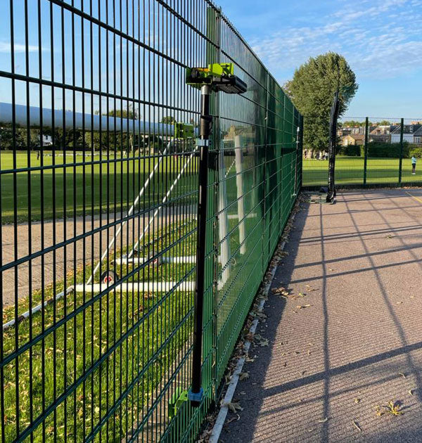 Close-up of Ritelite's Sports Lite Fence Mast Adapter mounting a camera or sensor on a green wire fence between a grassy field and paved path, with soccer goals and trees visible in the background under a blue sky.
