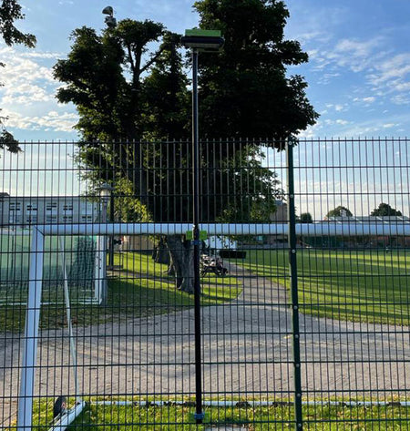 A Ritelite Sports Lite Fence Mast Adapter stands directly behind a metal fence on a sports field, perfectly aligned to appear as part of the goalpost. Trees and a building are visible in the partly cloudy background.