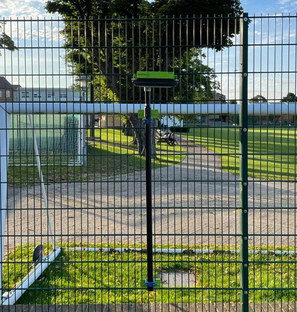 A Ritelite Sports Lite Fence Mast Adapter is attached vertically to a sports fencing wire fence by a sports field, with green grass, trees, and a blue sky in the background.