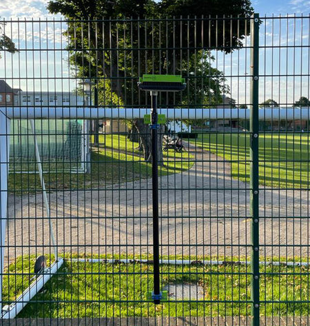 A Ritelite Sports Lite Fence Mast Adapter is attached vertically to a sports fencing wire fence by a sports field, with green grass, trees, and a blue sky in the background.
