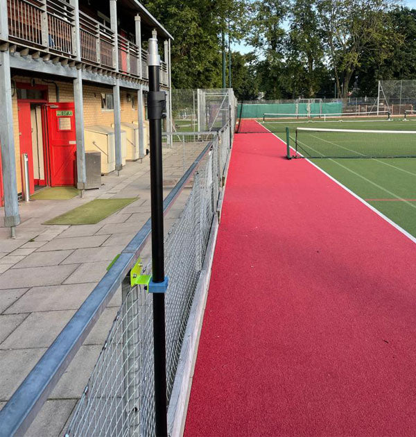 A red rubber running track runs beside a fenced sports field featuring Ritelite’s Sports Lite Fence Mast Adapter, with a building boasting red doors and a balcony nearby, plus trees and extra courts visible in the background.