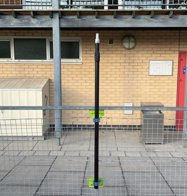 A Ritelite Sports Lite Fence Mast Adapter with a tall black adjustable mast and green brackets stands on paving in front of a brick building, ideal for sports fencing installations.