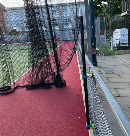 A black sports net covers a red and green court near a metal fence fitted with the Ritelite Sports Lite Fence Mast Adapter. A white van is parked by trees to the right, with a building visible in the background.