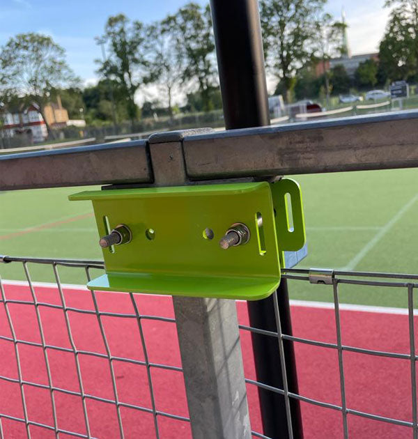 A close-up of the Ritelite Sports Lite Fence Mast Adapter, a green metal bracket secured with bolts, installed on a fence overlooking a sports field with a red track, green grass, trees, and buildings in the background.