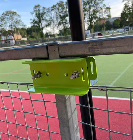 A close-up of the Ritelite Sports Lite Fence Mast Adapter, a green metal bracket secured with bolts, installed on a fence overlooking a sports field with a red track, green grass, trees, and buildings in the background.