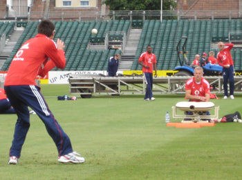 Several cricketers in red jerseys practice fielding on a grassy field. One operates a Bola Fielding Training Machine by Bola as another gets ready to catch the ball. The scene includes empty stadium seats and visible equipment in the background.