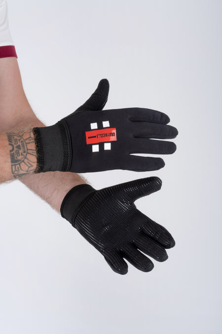 A person with a tattooed forearm models black Gray Nicolls Pro Catching Gloves. The top glove displays a red logo, while the palm of the other has a textured, non-slip surface. The background is light gray.