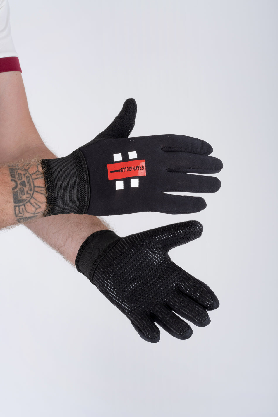 A person with a tattooed forearm models black Gray Nicolls Pro Catching Gloves. The top glove displays a red logo, while the palm of the other has a textured, non-slip surface. The background is light gray.