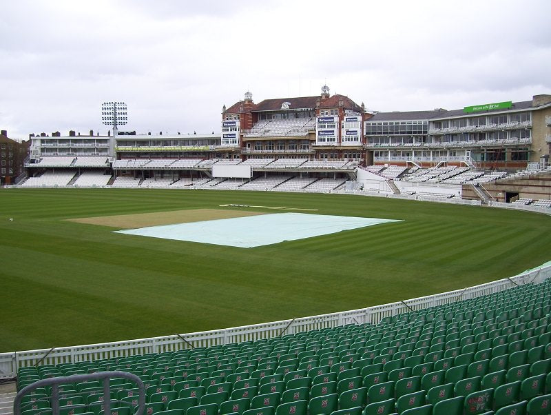A cricket stadium features rows of green seats with a Stuart Canvas Flatlay Lightweight Pitch Cover (25m x 4m) protecting the pitch center, while empty stands and buildings stand in the background under a cloudy sky.
