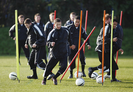 A group of young soccer players in black tracksuits practice dribbling around colorful Precision Training Flexi Boundary Poles & Bag on a grassy field, focusing on lateral maneuvering and active participation.