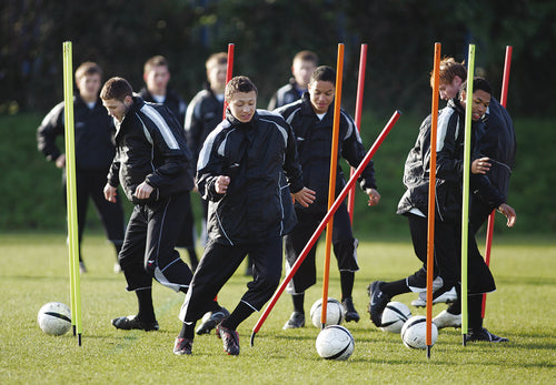 A group of young soccer players in black tracksuits practice dribbling around colorful Precision Training Flexi Boundary Poles & Bag on a grassy field, focusing on lateral maneuvering and active participation.