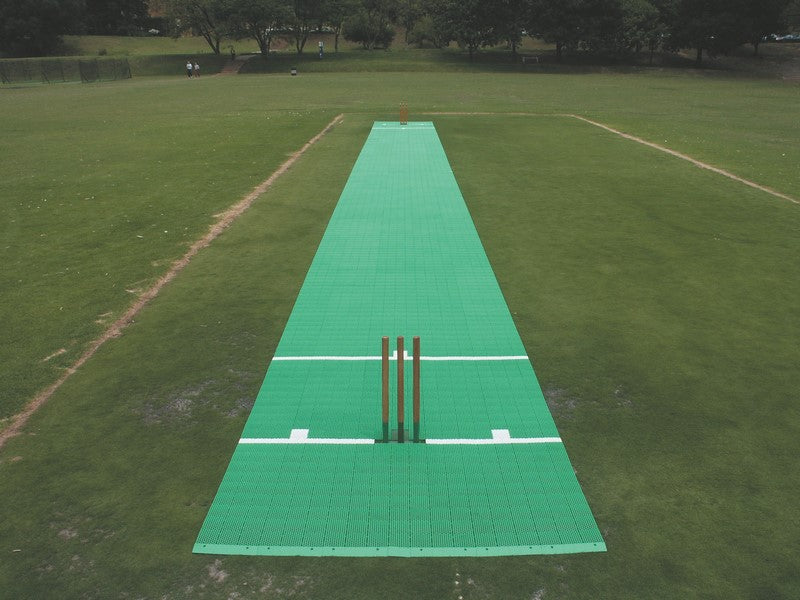 A 2G Flicx Match Pitch from Flicx UK is laid out on a grassy field, complete with stumps and white crease markings. Trees and fences can be seen in the background.