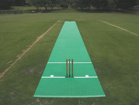 A 2G Flicx Match Pitch from Flicx UK is laid out on a grassy field, complete with stumps and white crease markings. Trees and fences can be seen in the background.