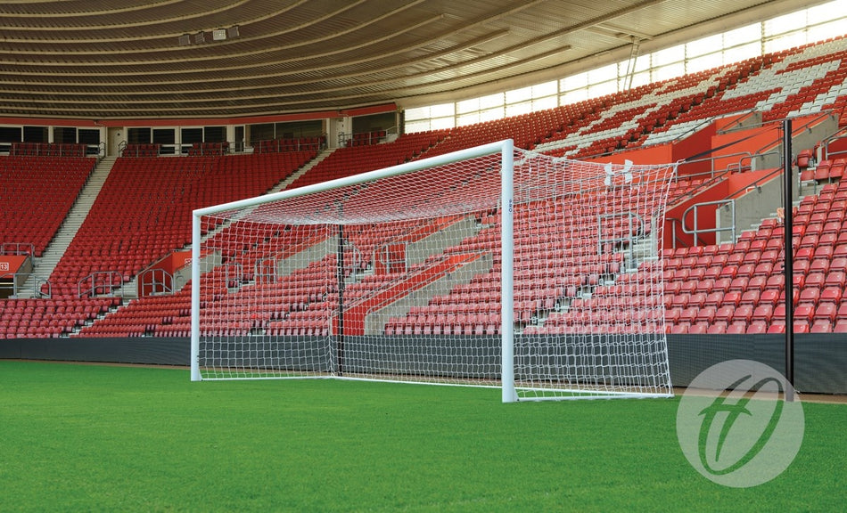 A Harrod Sport Stadium Pro Freehanging Net Supports - 4 Poles soccer goal stands on a green field in an empty stadium with red seats and a partially covered roof.