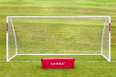 A Samba Sports Match Goal 9V9 16 x 7 stands on green grass with a red “samba” bag in front. A gently sloping grassy field is in the background, highlighting this durable uPVC football goal.