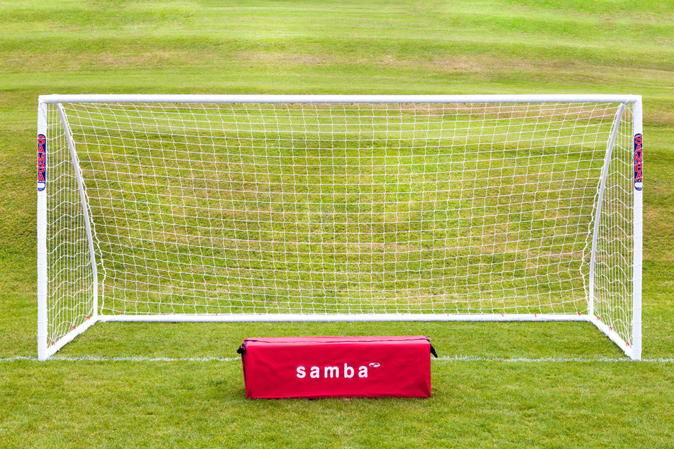A Samba Sports Match Goal 9V9 16 x 7 stands on green grass with a red “samba” bag in front. A gently sloping grassy field is in the background, highlighting this durable uPVC football goal.
