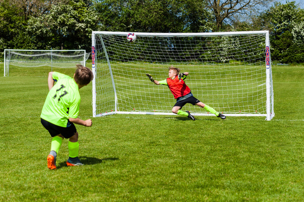 A young soccer player in a neon green jersey kicks a ball toward a Samba Sports Samba Match Goal 9V9 16 x 7 as a goalkeeper in red and black dives to make a save during a game on the grassy field.