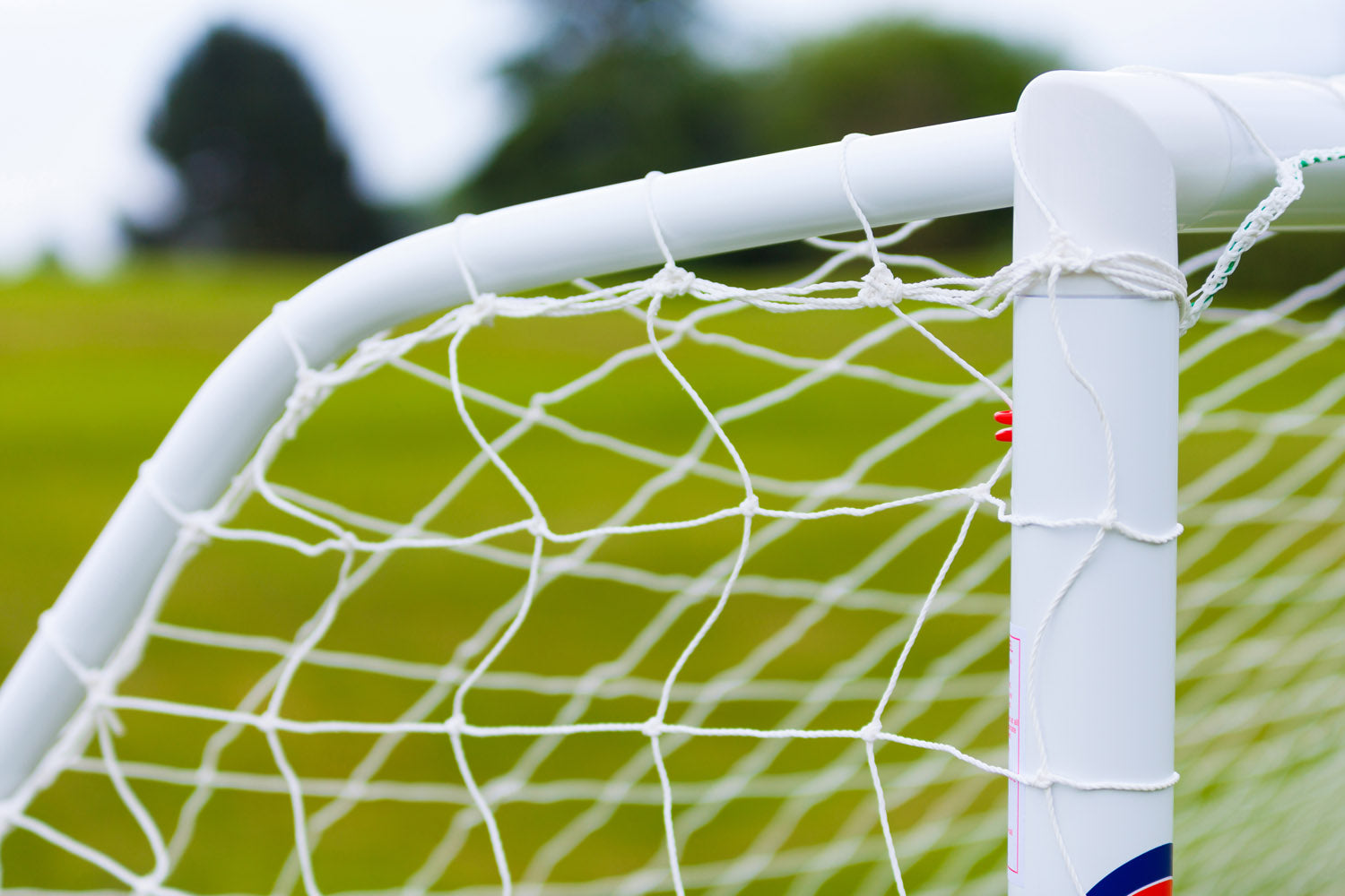 Close-up of a white Samba Sports Samba Match Goal 8 x 6, made from high-impact uPVC, with netting on green grass and blurred trees in the background.
