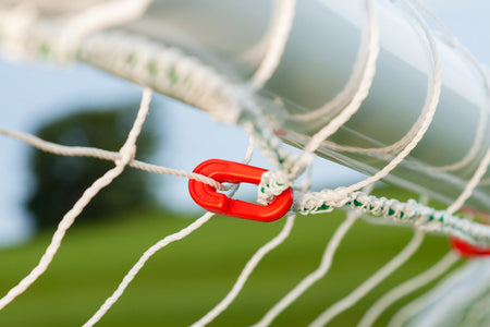 Close-up of a red plastic clip fastening the net to the high-impact uPVC frame of a Samba Sports Samba Match Goal 8 x 6, with green grass and a blurred tree in the background.